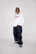 A model wearing a white long-sleeve crewneck sweatshirt with a red and black 'I LOVE NY' graphic, paired with black baggy pants featuring white flame embroidery near the cuffs, and black and white sneakers, set against a plain light gray studio background.