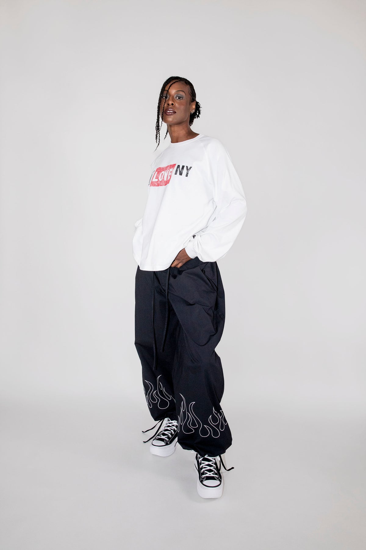 A model wearing a white long-sleeve crewneck sweatshirt with a red and black 'I LOVE NY' graphic, paired with black baggy pants featuring white flame embroidery near the cuffs, and black and white sneakers, set against a plain light gray studio background.