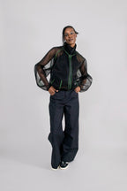 A model stands against a light gray studio background wearing a black sheer mesh zip-up jacket with green piping over a black top, paired with wide-leg dark trousers and black sneakers.