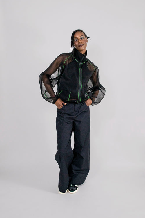 A model stands against a light gray studio background wearing a black sheer mesh zip-up jacket with green piping over a black top, paired with wide-leg dark trousers and black sneakers.