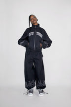 A model wearing a black zip-up track jacket and matching wide-leg pants set with white embroidery, including 'NEW YORK' on the chest and flame motifs near the ankles, paired with black and white sneakers against a light gray studio background.