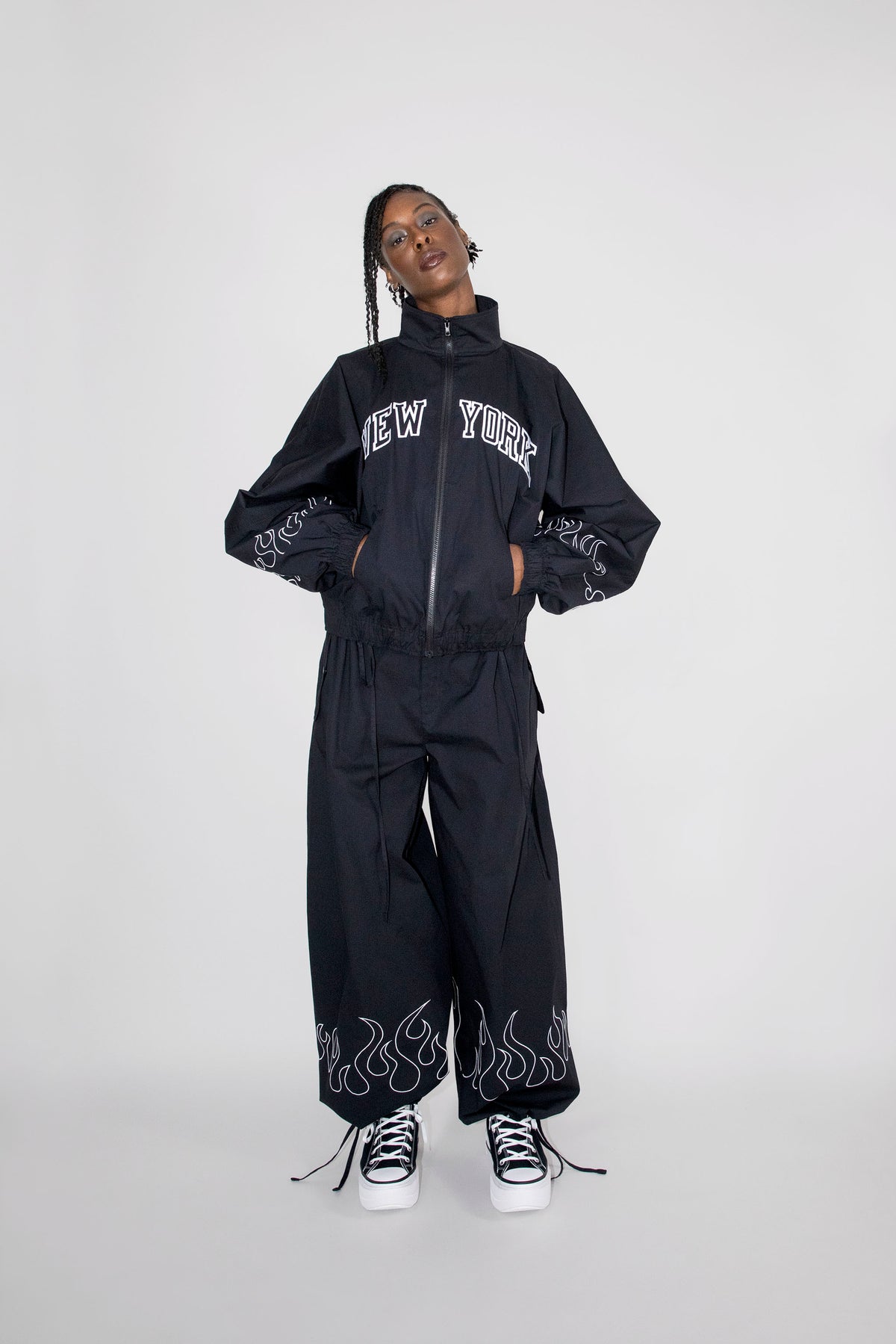 A model wearing a black zip-up track jacket and matching wide-leg pants set with white embroidery, including 'NEW YORK' on the chest and flame motifs near the ankles, paired with black and white sneakers against a light gray studio background.