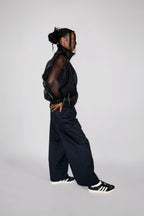 A model in profile wearing a black sheer mesh top, black wide-leg trousers, and black and white sneakers, standing against a light gray studio background.