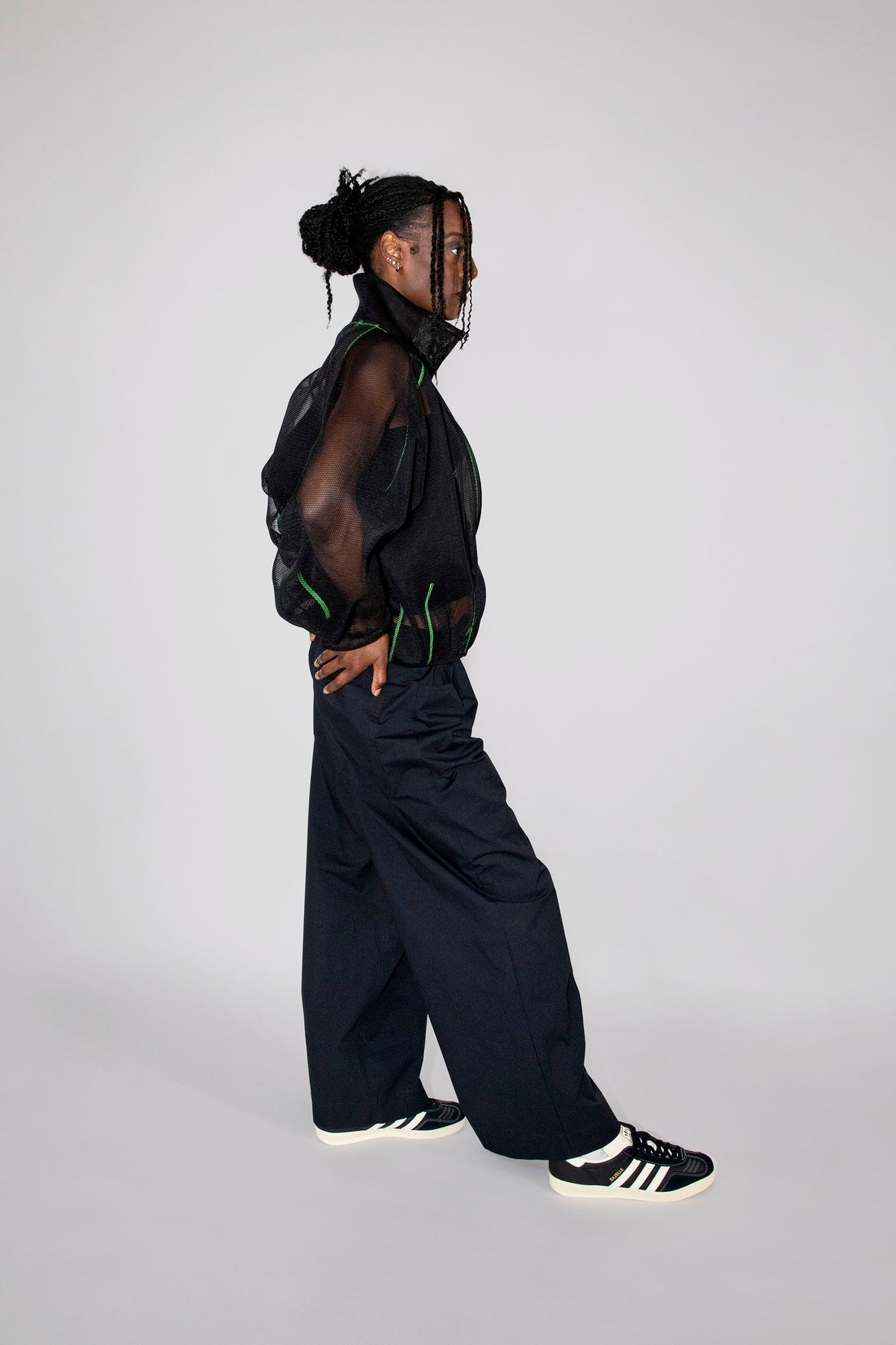A model in profile wearing a black sheer mesh top, black wide-leg trousers, and black and white sneakers, standing against a light gray studio background.
