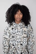 A person with voluminous black curly hair wearing a high-neck, long-sleeve trenchcoat with a black and white abstract organic pattern, photographed against a plain light gray background.