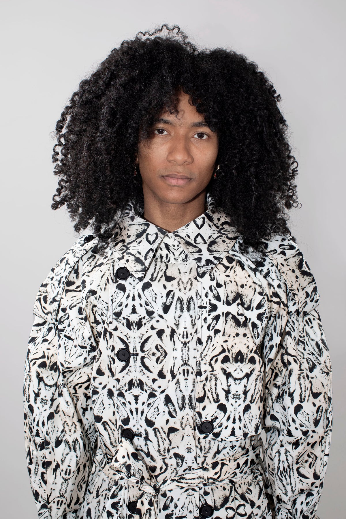 A person with voluminous black curly hair wearing a high-neck, long-sleeve trenchcoat with a black and white abstract organic pattern, photographed against a plain light gray background.