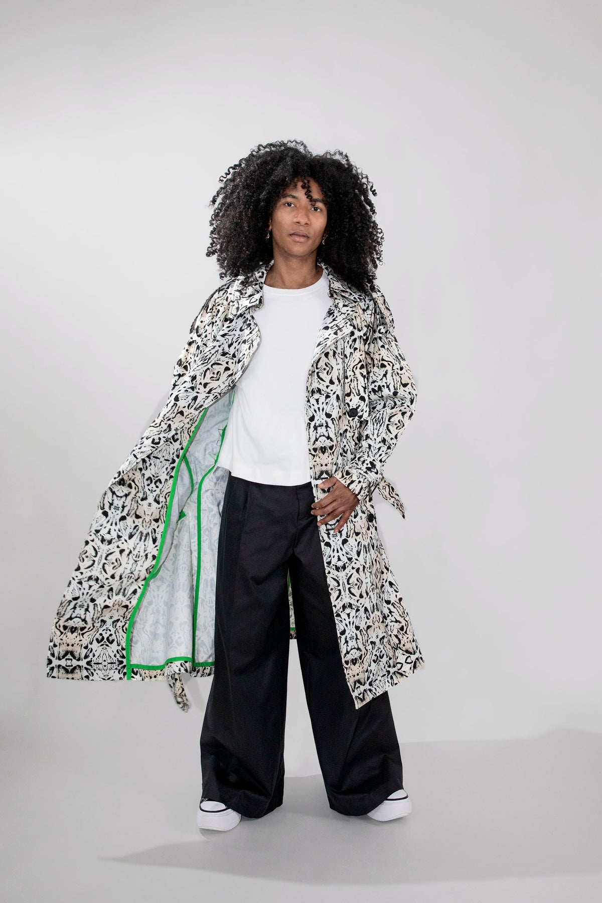 A model with curly black hair stands against a light gray studio background wearing a long white trenchcoat with a black abstract pattern, a white t-shirt, wide-leg black pants, and white sneakers.