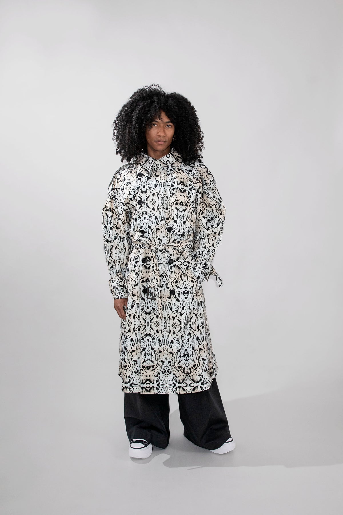 A model with curly black hair stands against a light gray studio background wearing a long-sleeved, high-neck patterned trenchcoat over black wide-leg pants and white sneakers.