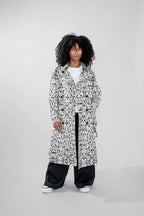 A model with curly black hair stands against a light gray studio background wearing a long white trenchcoat covered in a black abstract pattern, a white t-shirt, loose black pants, and white sneakers.