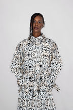 A model wearing a long-sleeved, collared trenchcoat with a black and white abstract pattern, standing against a plain light gray background.