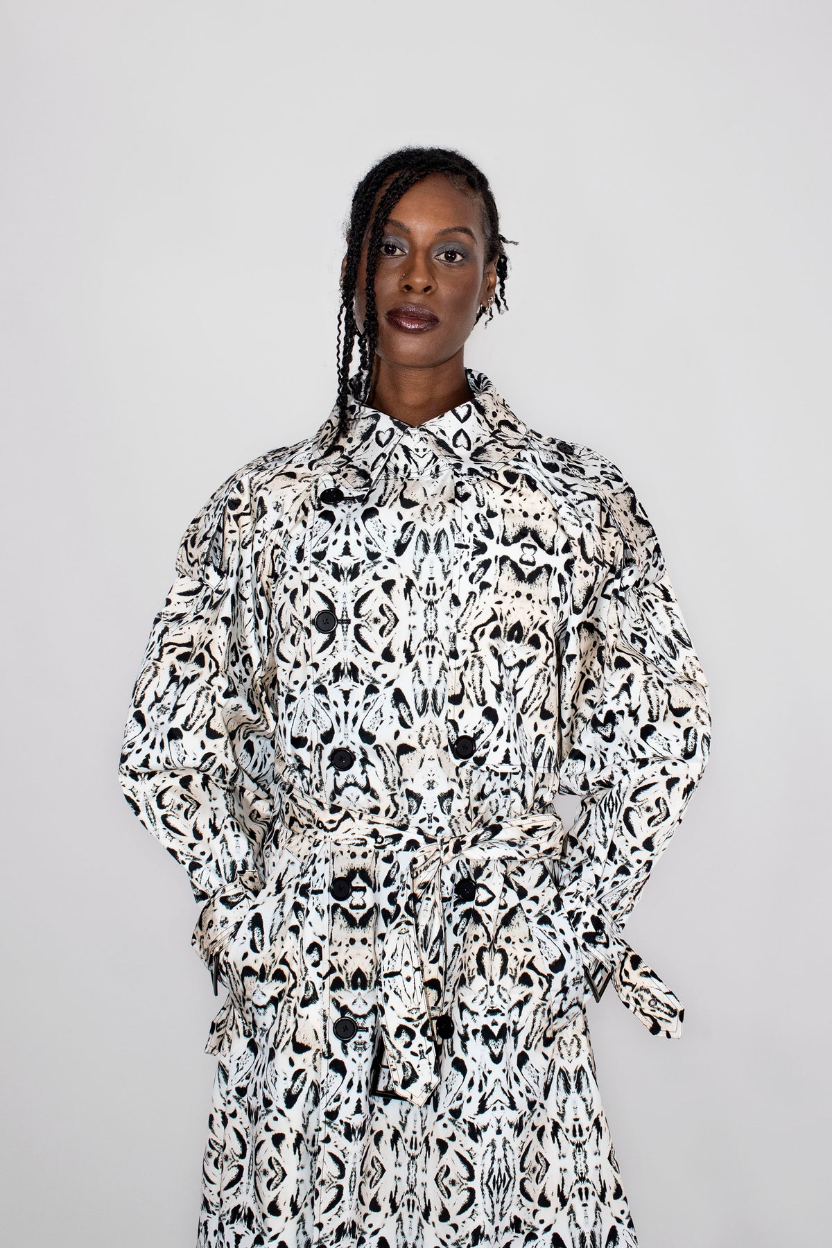 A model wearing a long-sleeved, collared trenchcoat with a black and white abstract pattern, standing against a plain light gray background.