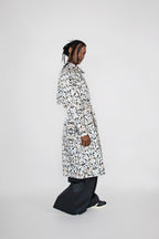 A model wearing a long-sleeved black and white abstract patterned trenchcoat over black wide-leg trousers, standing in profile against a light gray studio background.
