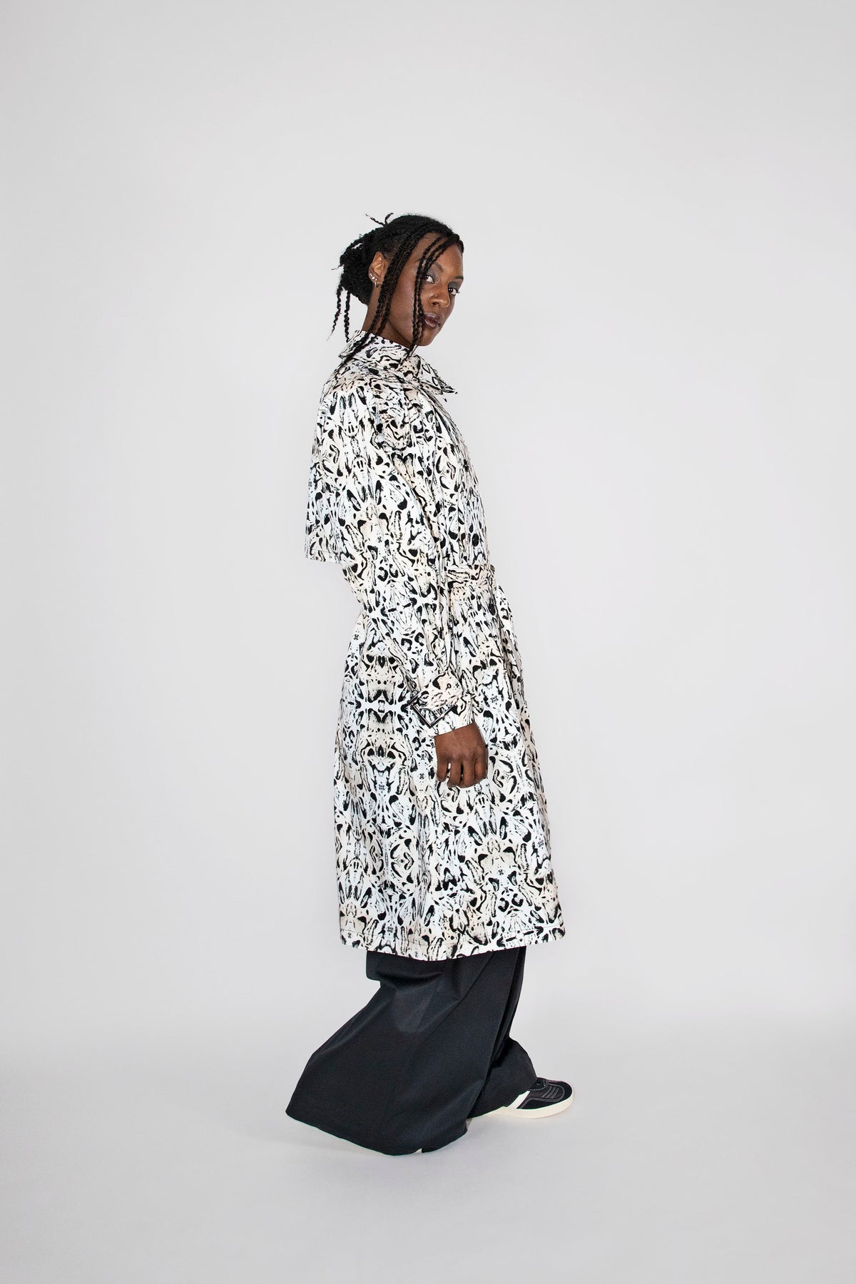 A model wearing a long-sleeved black and white abstract patterned trenchcoat over black wide-leg trousers, standing in profile against a light gray studio background.