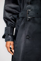 Close-up of a black trench coat worn by a model, showing the double-breasted front, belt buckle, and cuff details.