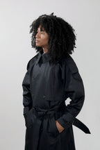 A person with voluminous curly black hair wearing a black double-breasted trench coat with a matching waist tie, standing against a light gray studio background.