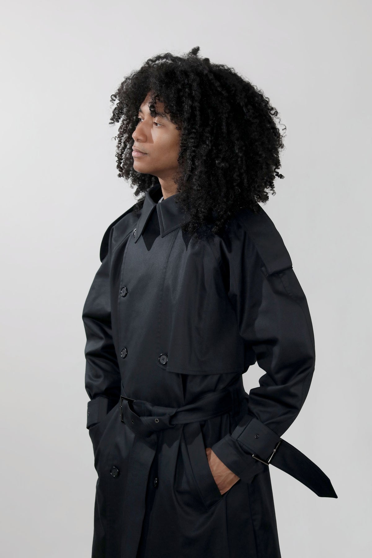 A person with voluminous curly black hair wearing a black double-breasted trench coat with a matching waist tie, standing against a light gray studio background.