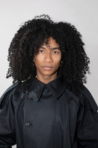 A person with voluminous curly black hair wearing a black collared trenchcoat photographed against a plain light gray studio background.