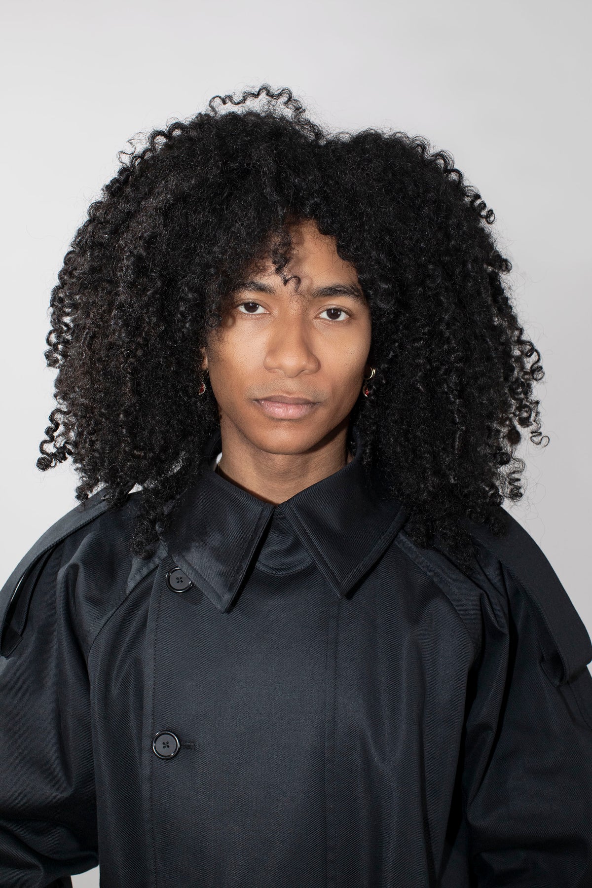 A person with voluminous curly black hair wearing a black collared trenchcoat photographed against a plain light gray studio background.