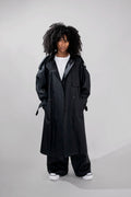 A model with curly black hair wearing a long black double-breasted trench coat over a white t-shirt, wide-leg black pants, and white sneakers, standing against a light gray studio background.