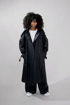 A model with curly black hair wearing a long black double-breasted trench coat over a white t-shirt, wide-leg black pants, and white sneakers, standing against a light gray studio background.