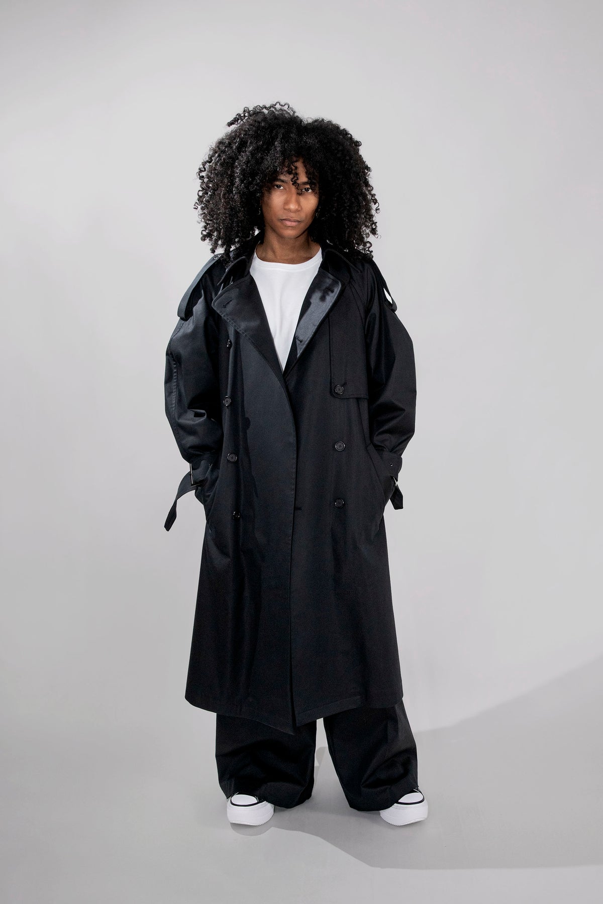 A model with curly black hair wearing a long black double-breasted trench coat over a white t-shirt, wide-leg black pants, and white sneakers, standing against a light gray studio background.