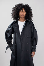 A model with voluminous black curly hair wearing a black double-breasted trench coat over a white t-shirt, photographed against a neutral light gray studio background.