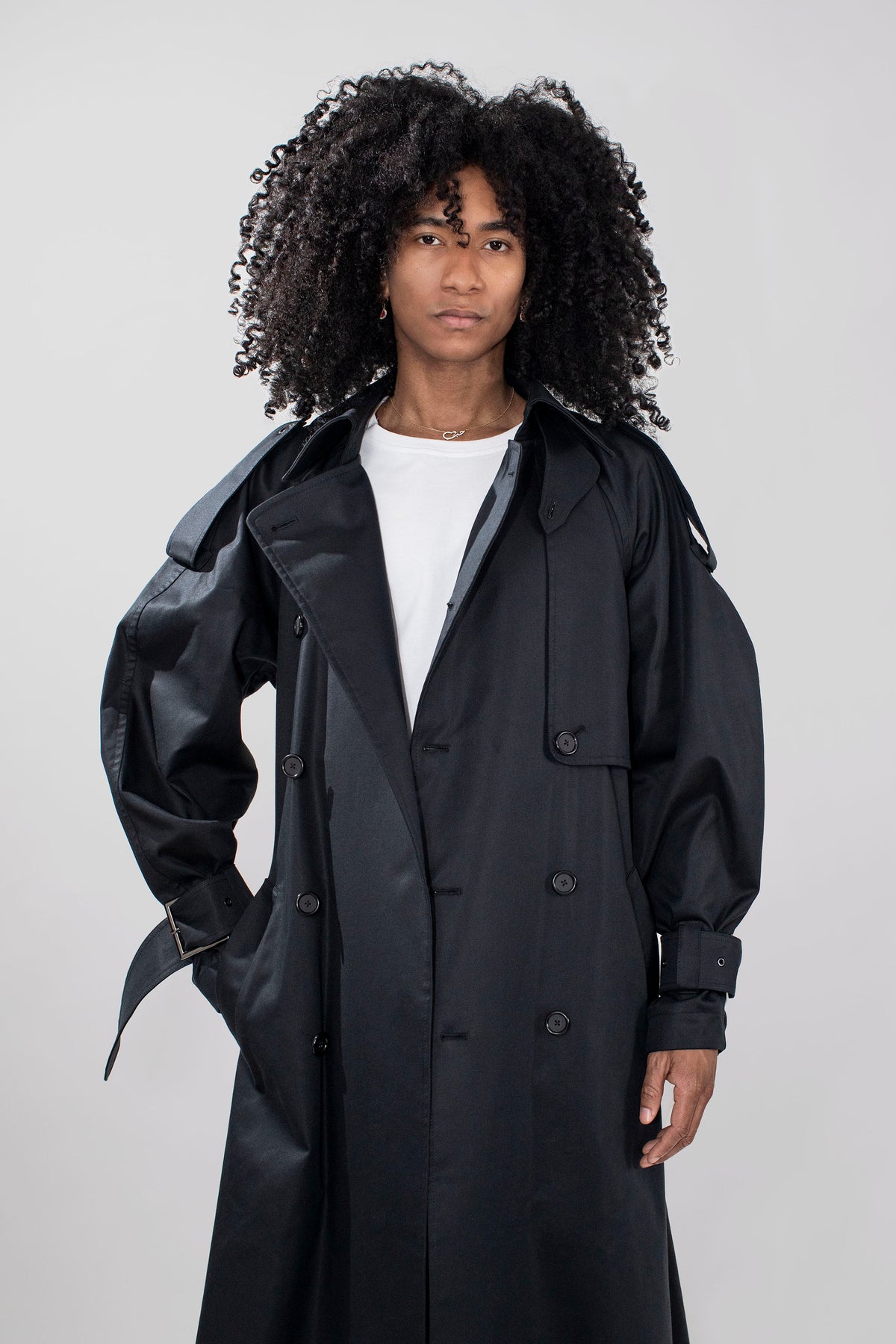 A model with voluminous black curly hair wearing a black double-breasted trench coat over a white t-shirt, photographed against a neutral light gray studio background.