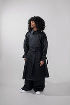 A model with curly black hair wearing a long black trench coat, wide-leg black pants, and white sneakers, standing against a light gray studio background.