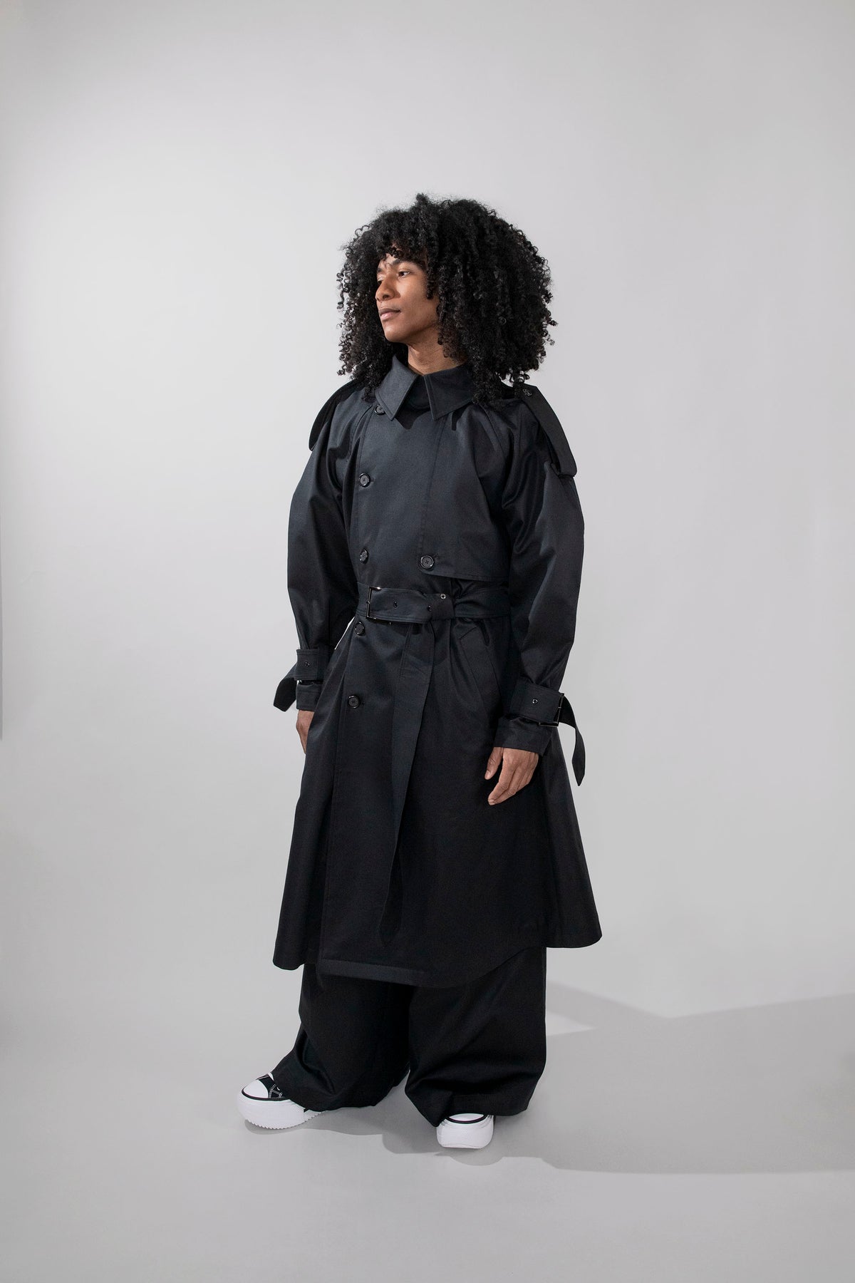 A model with curly black hair wearing a long black trench coat, wide-leg black pants, and white sneakers, standing against a light gray studio background.