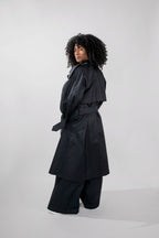 A model with curly black hair is shown from a side-back angle wearing a black trench coat with a matching belt and wide-leg black trousers, set against a plain light gray studio background.