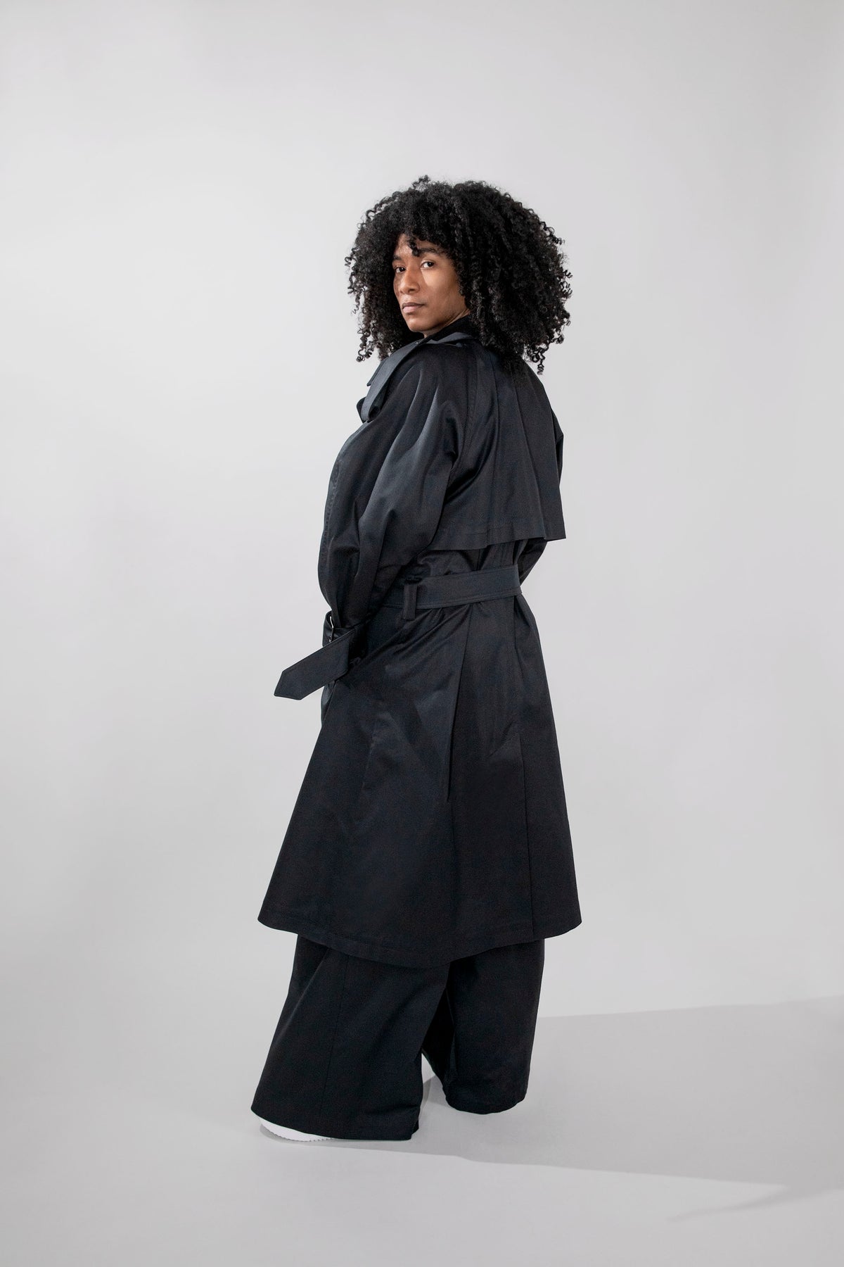 A model with curly black hair is shown from a side-back angle wearing a black trench coat with a matching belt and wide-leg black trousers, set against a plain light gray studio background.