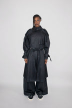 A model stands against a light gray studio background wearing a long black double-breasted trench coat with a waist tie, matching wide-leg pants, and black sneakers with white soles.