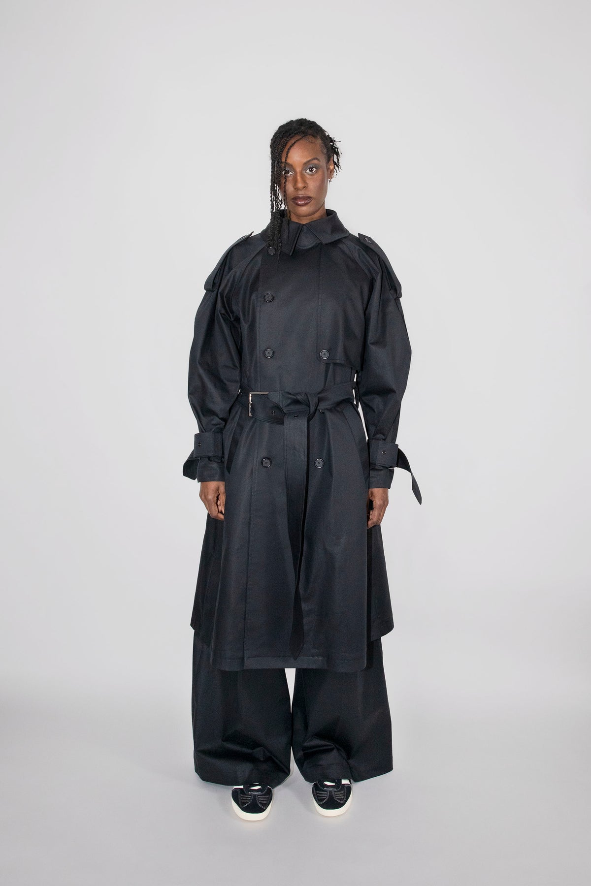 A model stands against a light gray studio background wearing a long black double-breasted trench coat with a waist tie, matching wide-leg pants, and black sneakers with white soles.