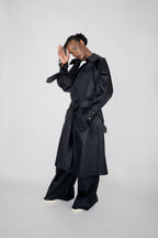 A model wearing a long black trench coat with a matching belt, wide-leg black trousers, and white sneakers, posing against a light gray studio background.