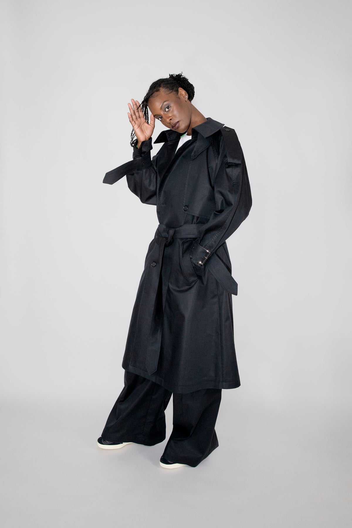A model wearing a long black trench coat with a matching belt, wide-leg black trousers, and white sneakers, posing against a light gray studio background.