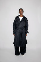 A model stands against a light gray studio background wearing a long black trench coat over a white top and black wide-leg pants.
