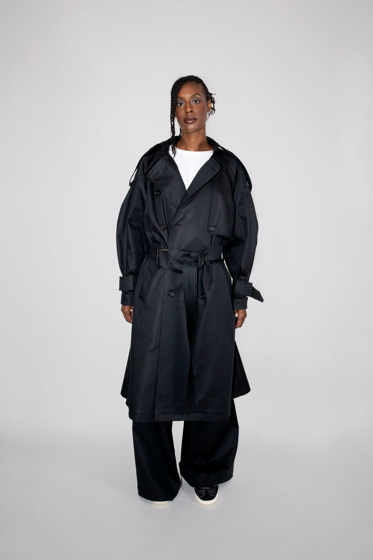 A model stands against a light gray studio background wearing a long black trench coat over a white top and black wide-leg pants.