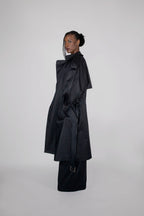 A model wearing a long black trench coat, photographed against a seamless light gray studio background.