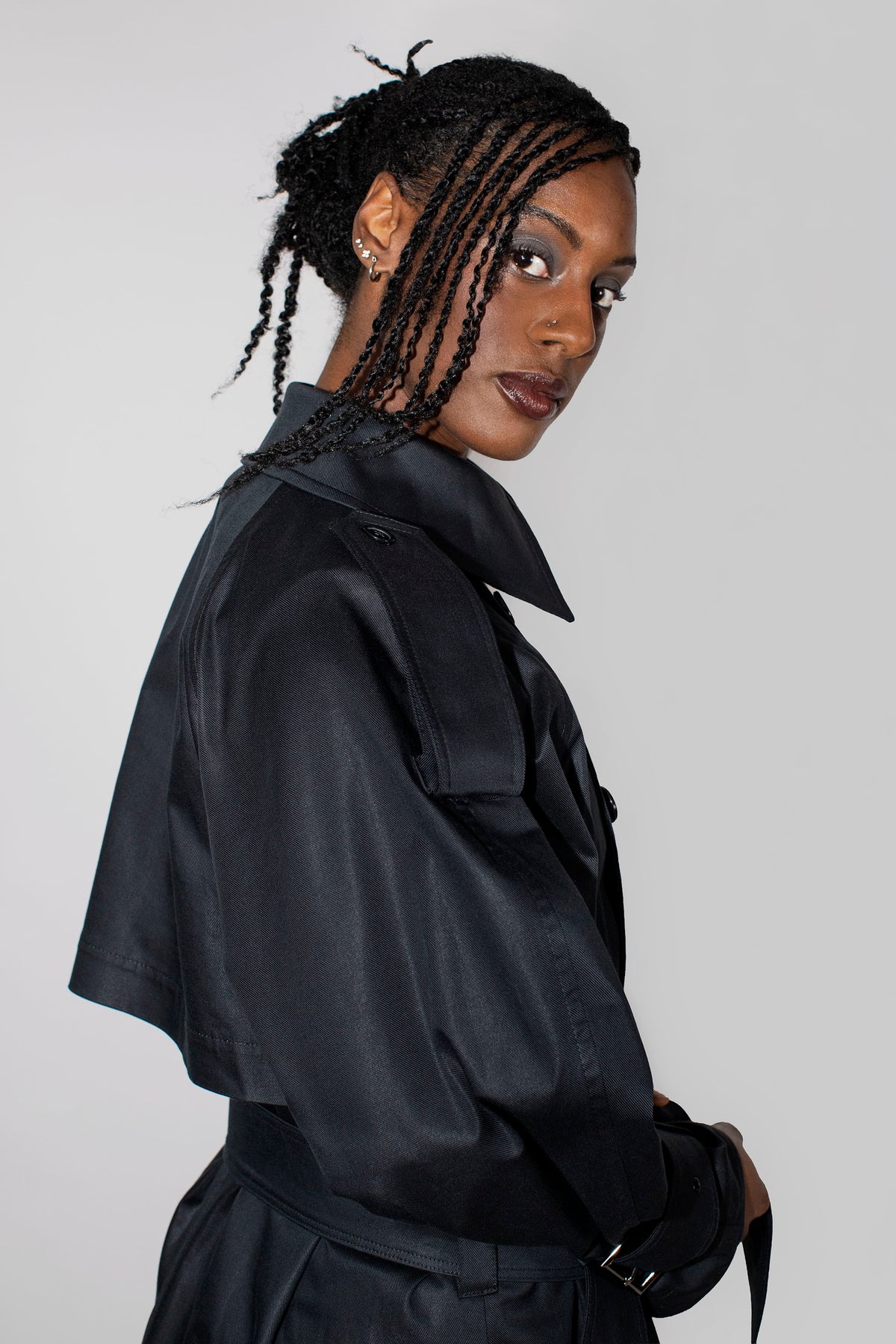 A woman with braided hair wearing a black trench coat, photographed against a plain light gray studio background.
