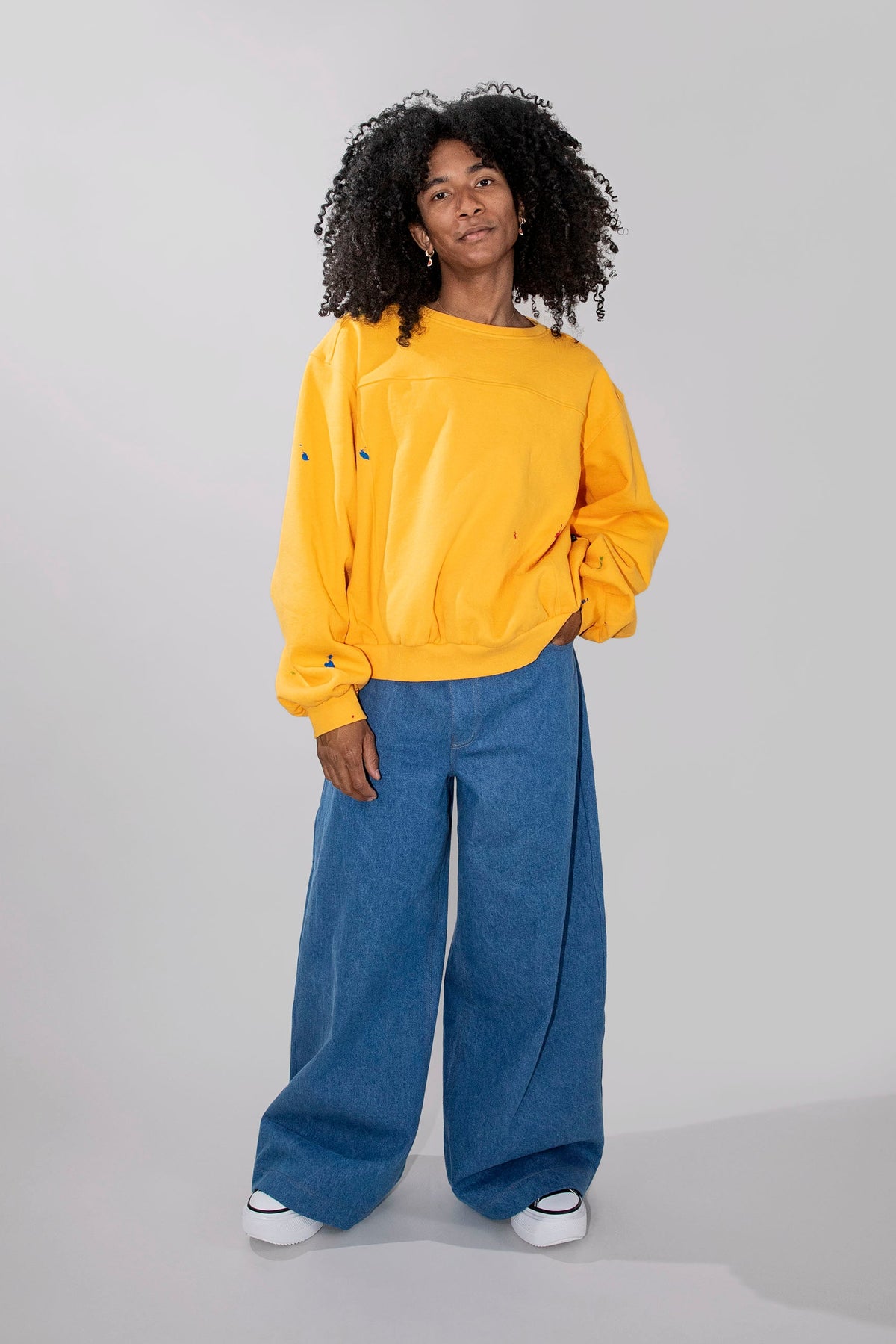 A model with curly black hair stands against a light gray studio background wearing a bright yellow oversized sweatshirt, wide-leg blue jeans, and white sneakers.