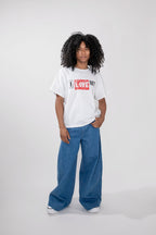 A model with curly black hair stands against a light gray studio background wearing a white graphic t-shirt with a red and black 'I LOVE NY' print, paired with wide-leg blue jeans and white sneakers.