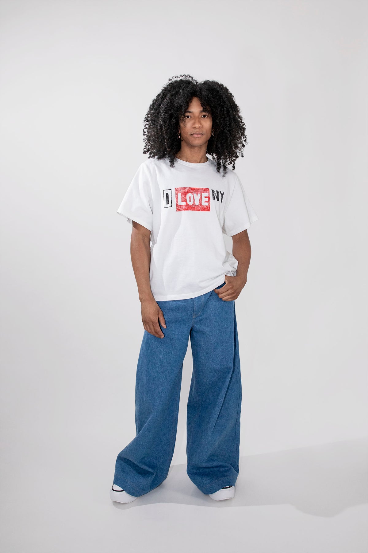A model with curly black hair stands against a light gray studio background wearing a white graphic t-shirt with a red and black 'I LOVE NY' print, paired with wide-leg blue jeans and white sneakers.