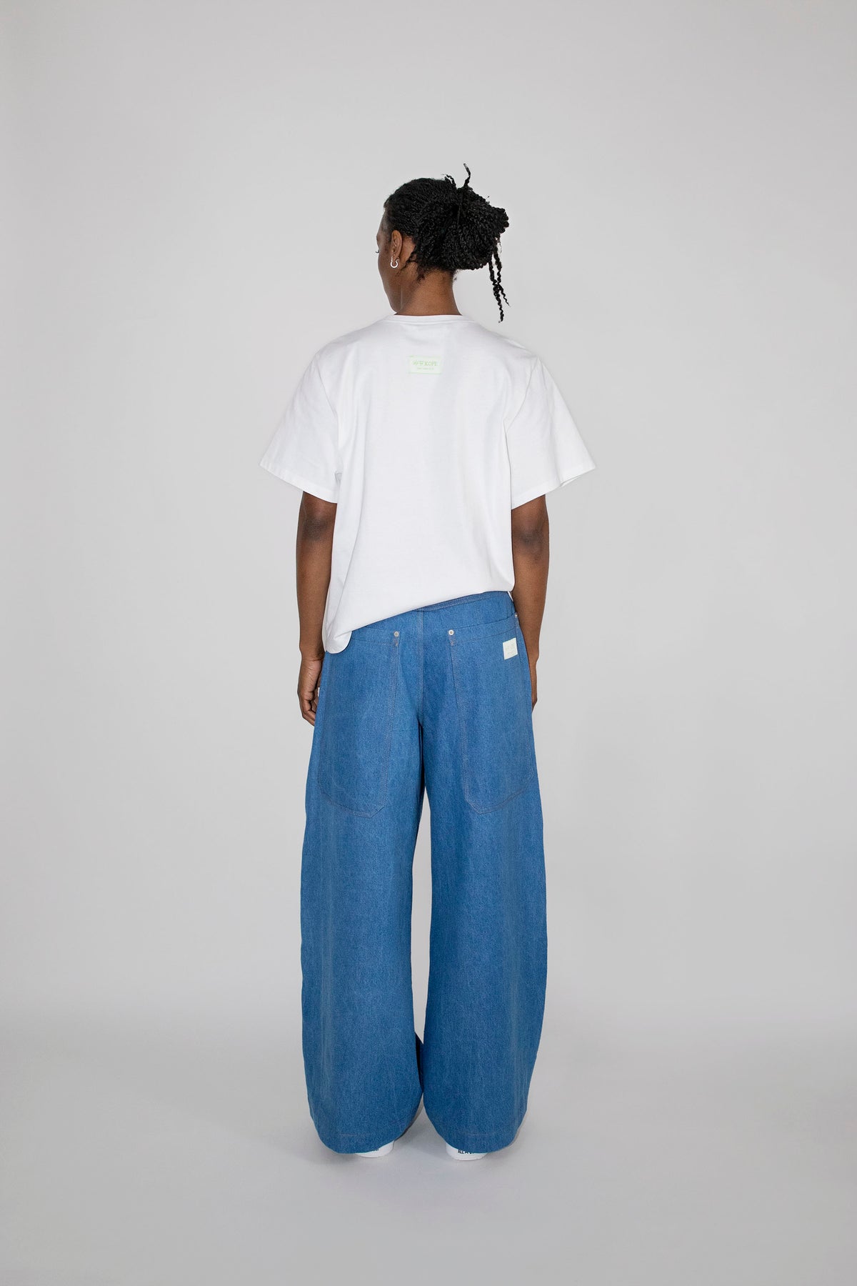 A person standing with their back to the camera, wearing a white short-sleeve t-shirt and wide-leg blue denim pants against a light gray studio background.