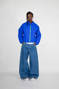 A model stands against a light gray studio background wearing a cobalt blue quilted zip-up jacket with a neon green zipper, wide-leg blue jeans, and white sneakers.