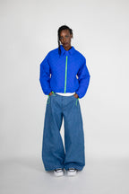 A model stands against a light gray studio background wearing a cobalt blue quilted zip-up jacket with a neon green zipper, wide-leg blue jeans, and white sneakers.