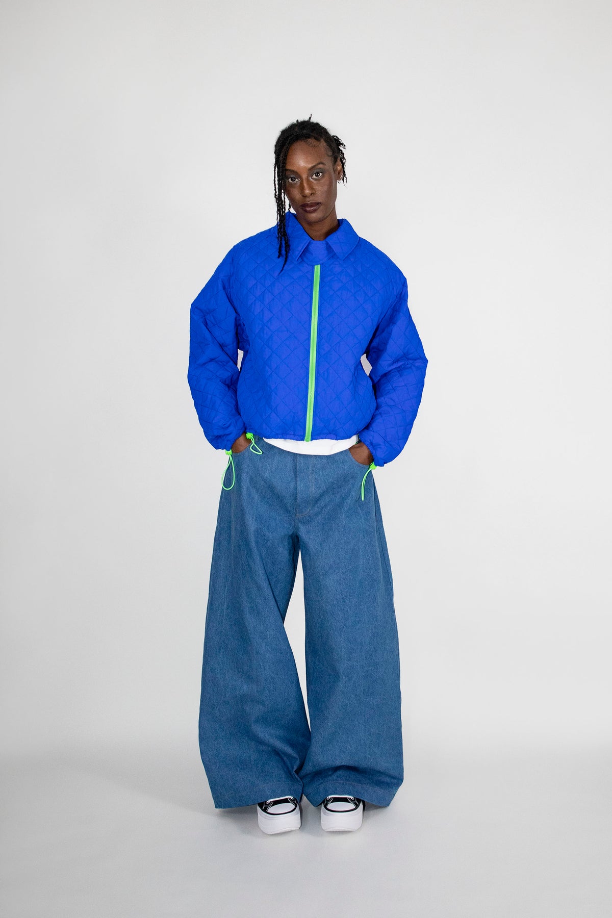 A model stands against a light gray studio background wearing a cobalt blue quilted zip-up jacket with a neon green zipper, wide-leg blue jeans, and white sneakers.