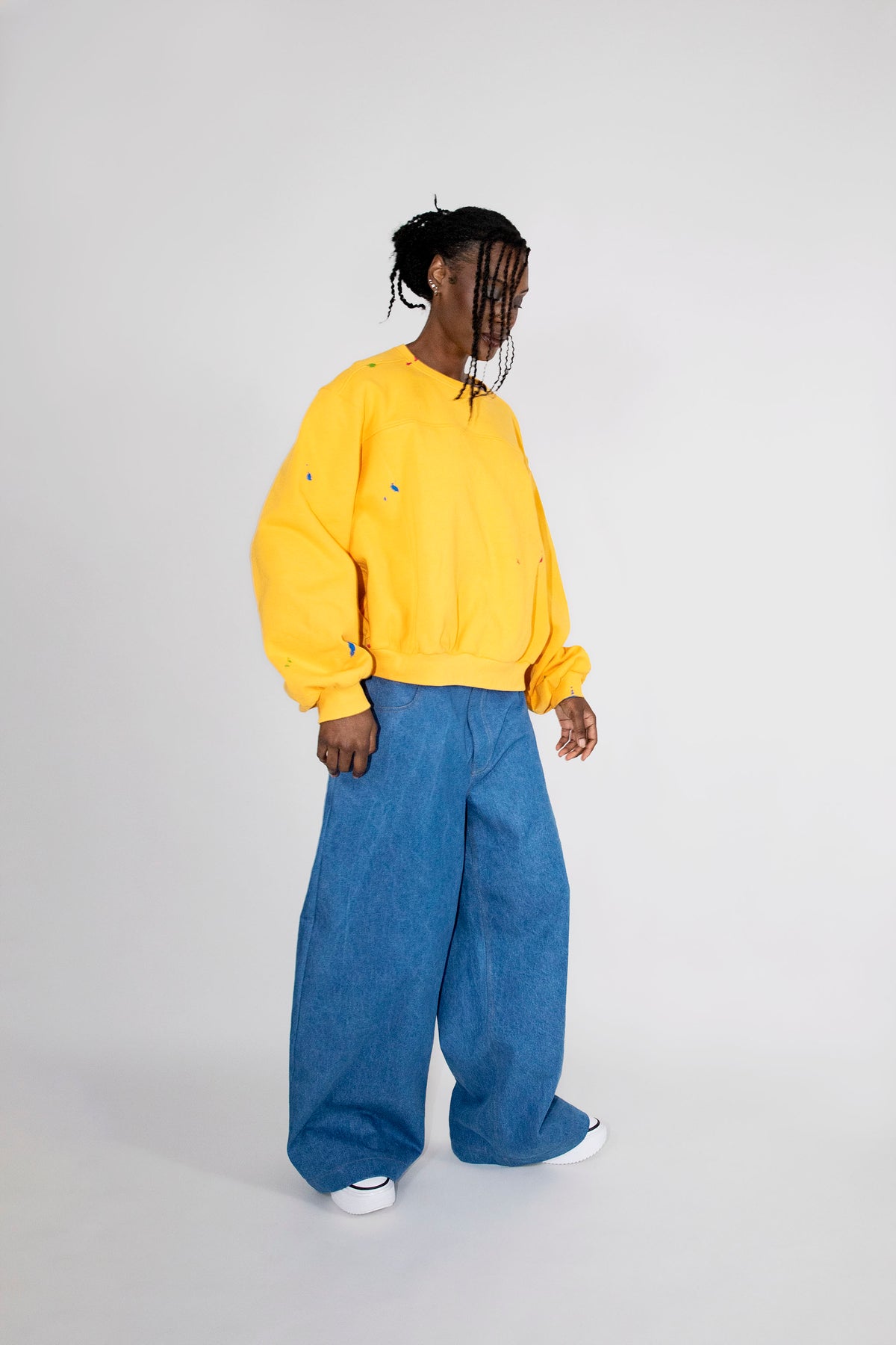 A model wearing a bright yellow oversized crewneck sweatshirt and wide-leg blue denim pants, standing against a plain light gray studio background.