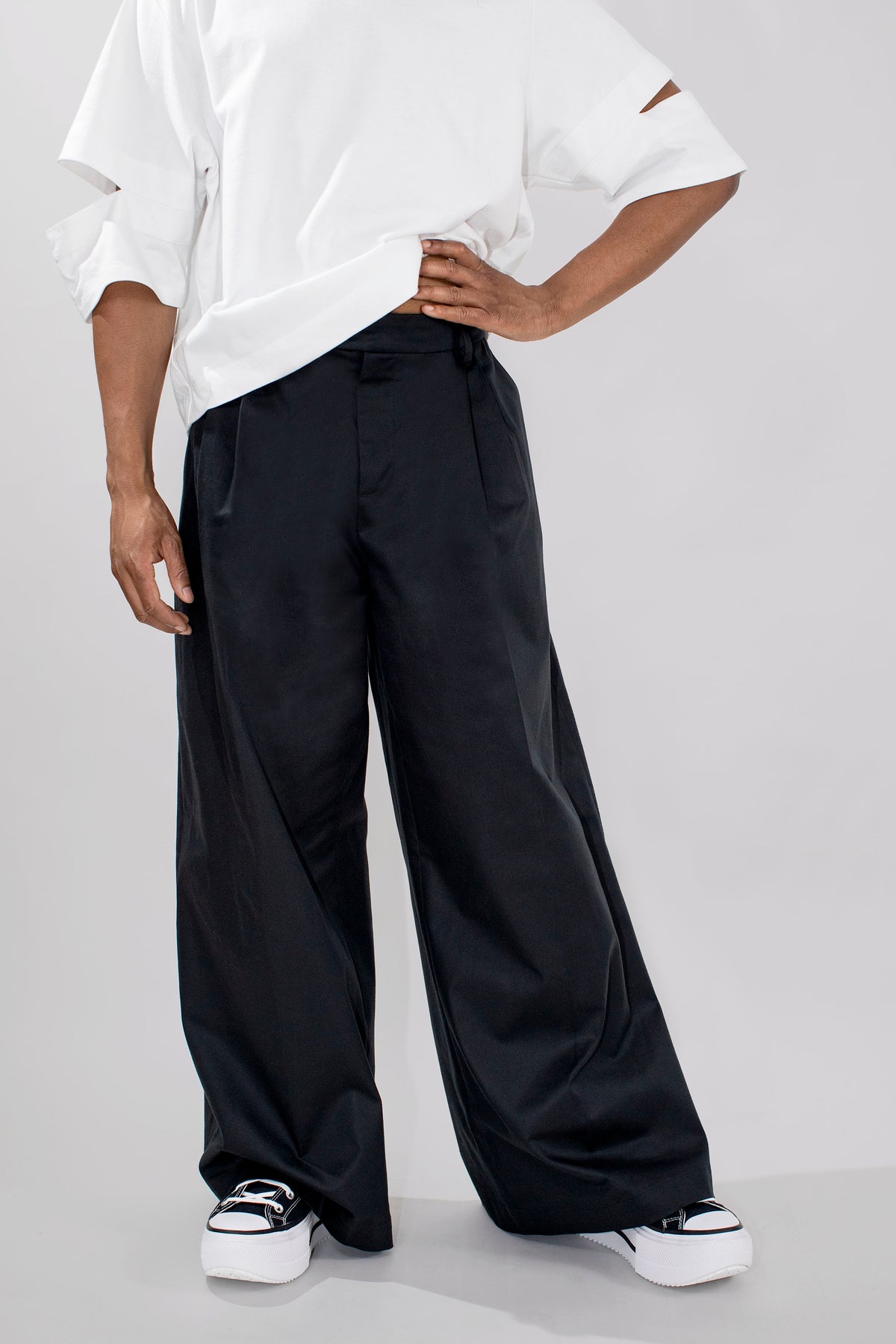A model wearing black wide-leg trousers with a white shirt and black-and-white platform sneakers, shown from the chest down against a light gray studio background.