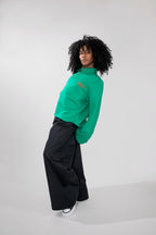 A model with curly black hair is shown from the side, wearing a vibrant green polo long-sleeve top with a small orange embroidered logo on the chest, paired with black wide-leg trousers and white sneakers, against a light gray studio background.