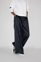 A model from the waist down wearing loose, wide-leg black trousers, a white t-shirt, and black sneakers with white stripes, standing against a light gray studio background.